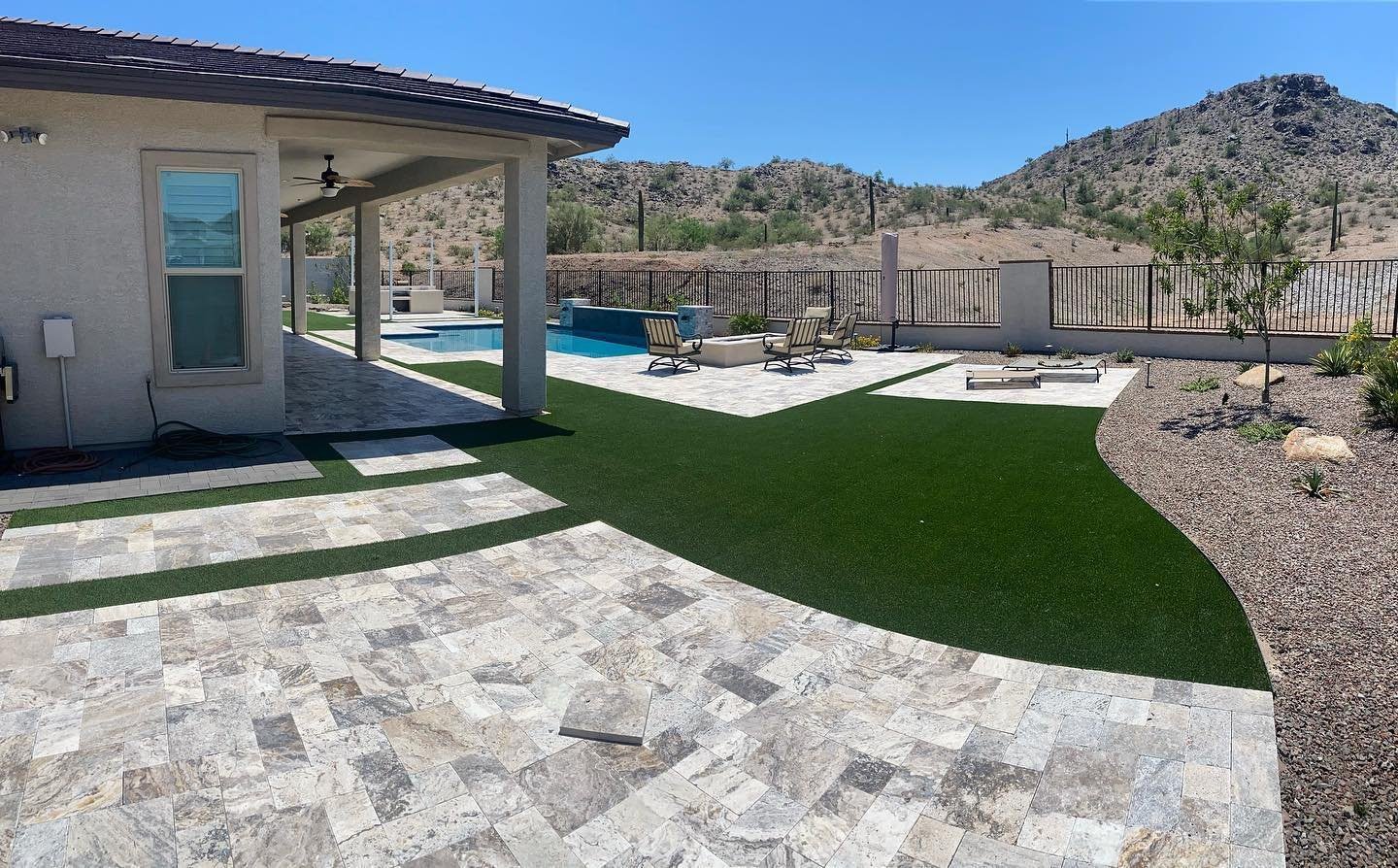 A backyard in Tucson, AZ, with a stone patio and a swimming pool adjacent to the house. The area features lounge chairs, a fire pit, and artificial turf by a skilled landscaping company. In the background, a mountainous landscape is enclosed by a fence. The house has large windows and a ceiling fan.