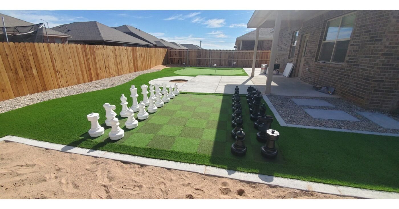 A backyard in Casas Adobes with a large chess set on Tucson Artificial Grass, flanked by a patio area and a putting green. The yard is enclosed by a wooden fence, and the patio is partially shaded by a roof extension from the house. Nearby houses are visible in the background, showcasing expert artificial grass installation.