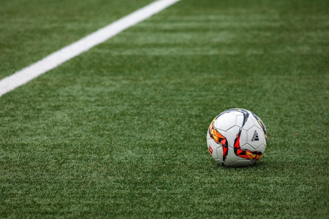 A soccer ball with a white, orange, and black design rests on a green, well-maintained astro turf sports field near a white boundary line.