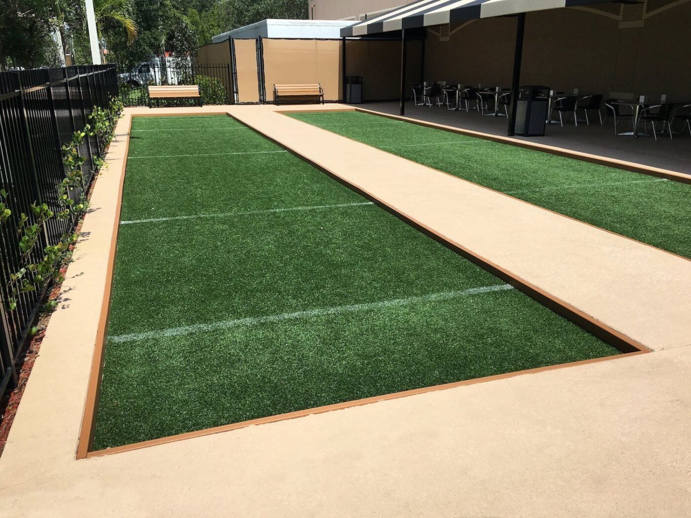 Two parallel bocce courts with Tucson Artificial Grass surfaces are bordered by beige walkways. The courts are fenced on one side, with seating areas and tables under a shaded structure in the background. Small plants line the fence. A wooden bench is visible.