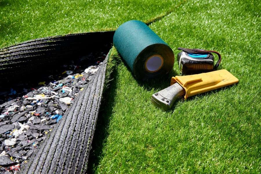 A section of artificial grass is being installed on a lawn. Part of the grass mat is lifted, revealing the underlying layer. Nearby, there is a roll of green tape, a utility knife in a yellow sheath, and a flashlight laid out on the grass. For expert pet turf installation in Tucson AZ, get your free quote today!