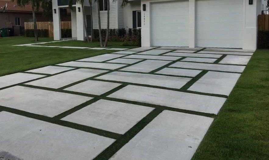 A modern driveway in Tucson AZ showcases a geometric design with large rectangular concrete slabs separated by strips of artificial grass. The driveway leads to a double garage with white doors. The surrounding lawn is neatly maintained, and a contemporary house is visible in the background.