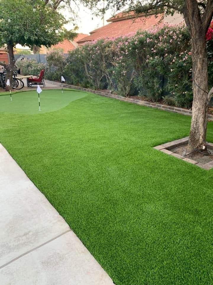 A backyard in Marana AZ features vibrant green artificial turf installation, a putting green with several holes and flags, a tree on the right, and a paved area on the left. Bicycles and a seating area are visible in the background near lush bushes and a wooden fence. The project is licensed & insured.