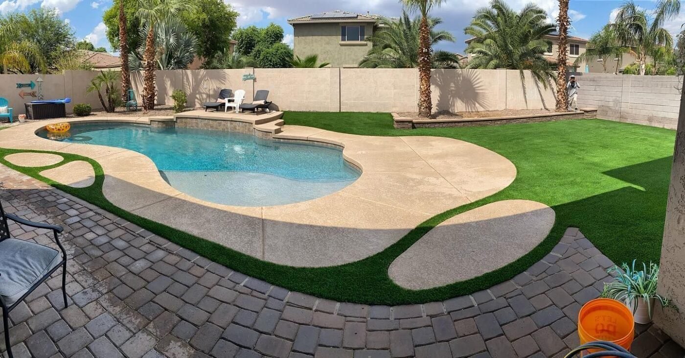 A backyard with a curvy swimming pool surrounded by a concrete patio and artificial grass installation. The area features lounge chairs near the pool and is enclosed by a tall, white fence. There are palm trees along the fence and a paver walkway in the foreground, perfect for Casas Adobes living.