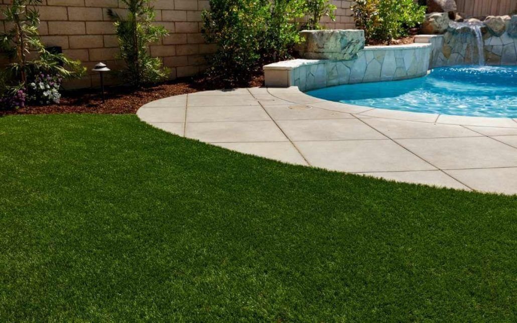 A backyard with a curved concrete patio seamlessly bordering a swimming pool. The pool features built-in stone seating areas and a waterfall. The patio is surrounded by lush artificial turf installation and landscaped with various plants and flowers against a brick wall, making it the perfect oasis in Marana, AZ.