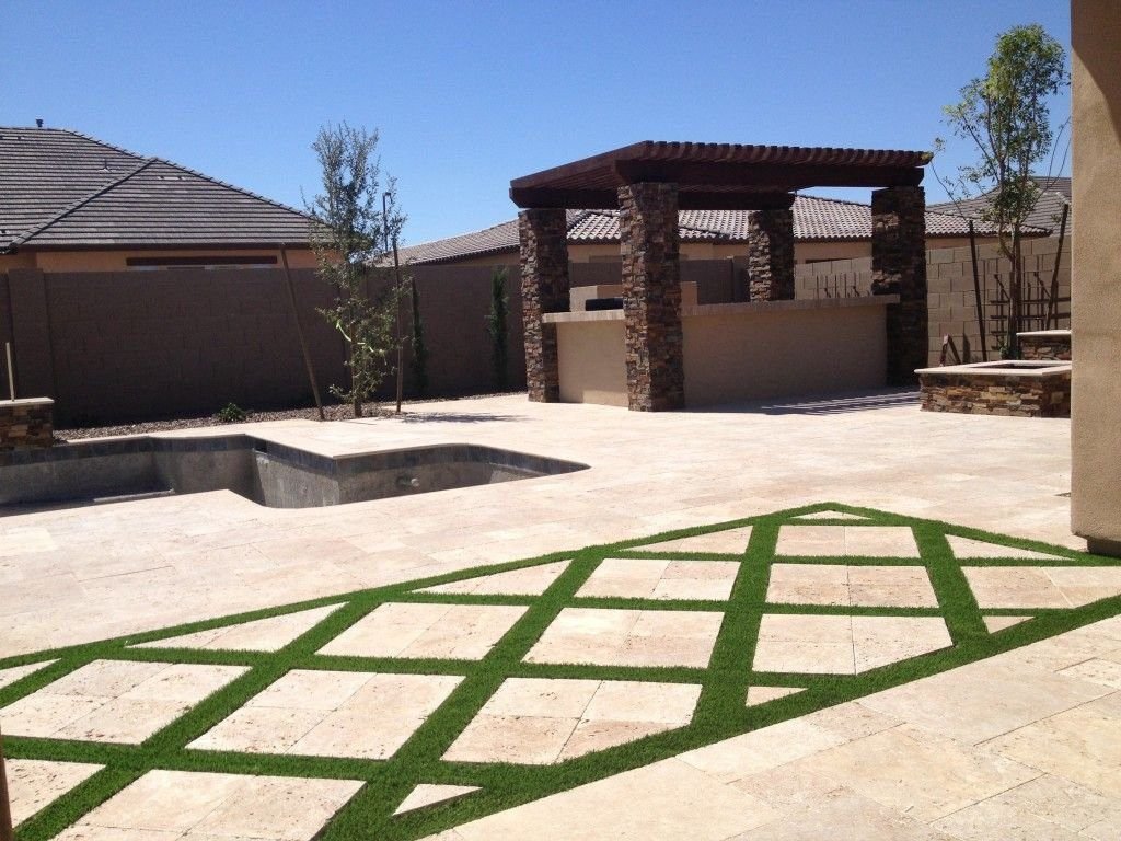 A backyard in Marana AZ features a pergola with a stone base, set next to an empty swimming pool. The patio boasts a stone tile design with green grass strips arranged in a diamond pattern, thanks to an artificial turf installation by a reliable company. The area is surrounded by a stone wall and houses with slanted roofs.